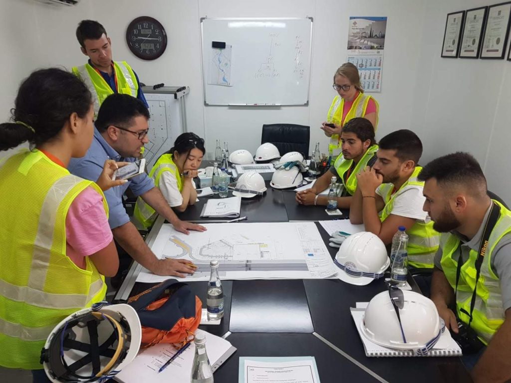 A group of students gather around a set of blueprints, which are spread out on a table. The students are wearing reflective vests, and their hard hats are sitting on the table.