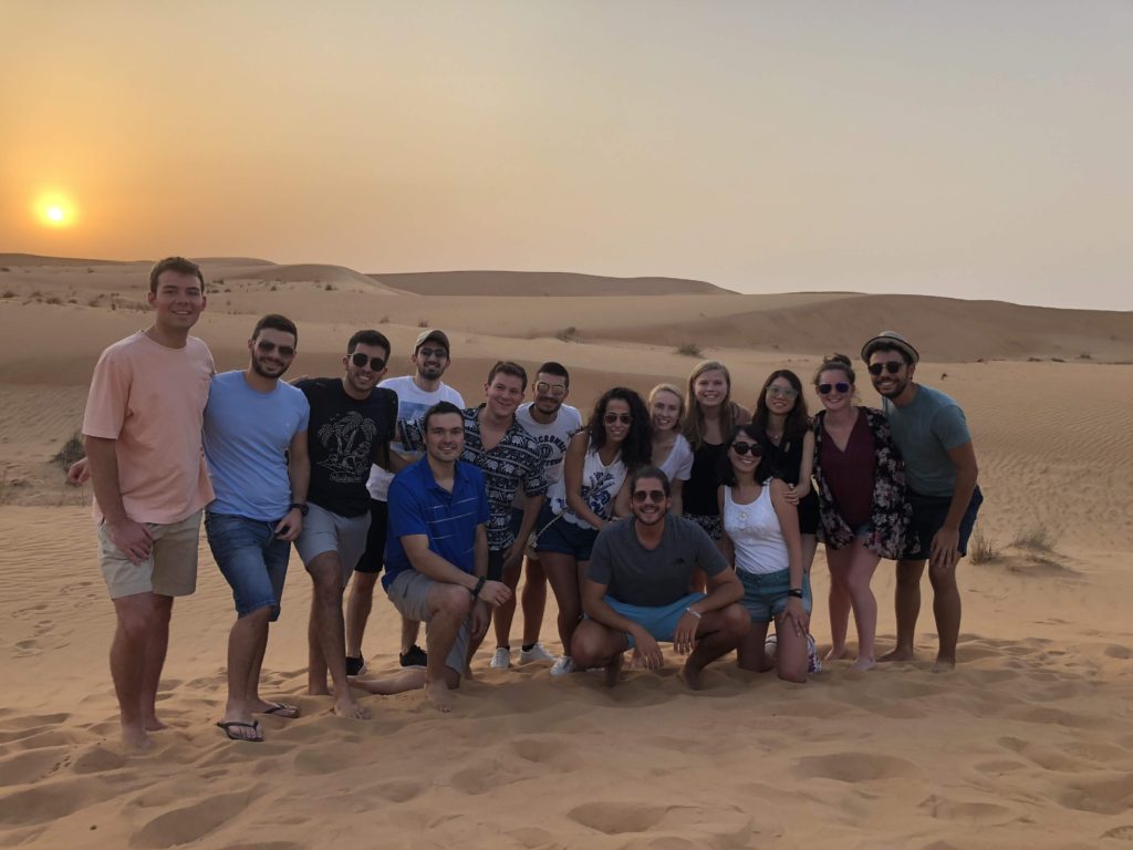 A group of fourteen students pose for a photo on a sand dune at sunset