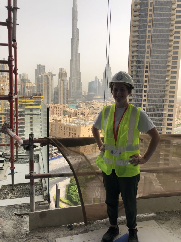 Picture of Haley standing at a construction site, with the city skyline behind her. She wears a reflective vest and hard hat.