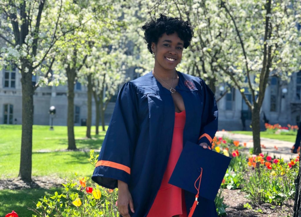 Simone dressed in her graduation gown, holding her graduation cap, with the hall of languages and flowering trees in the background